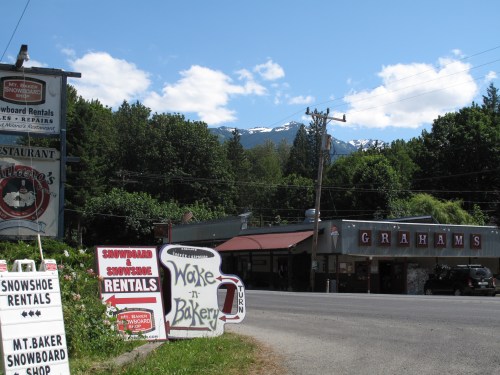 A roadside view featuring a snowboard rental shop, a bakery, and mountains in the background on a sunny day.