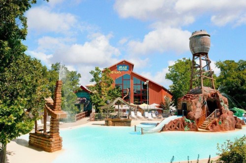 A water park featuring a large pool, water slides, and a rustic building in the background surrounded by trees.