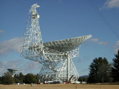 A large white radio telescope structure stands against a blue sky, surrounded by trees and a smaller dish in the background.