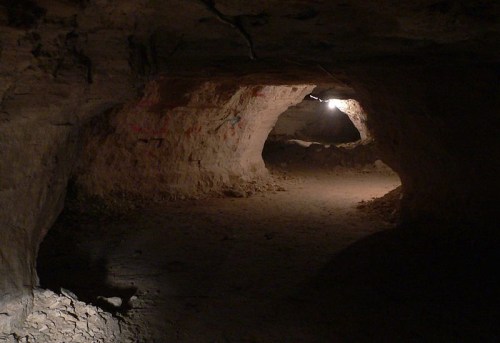 A dimly lit cave with rough stone walls and a sandy floor, featuring two arching openings in the distance.