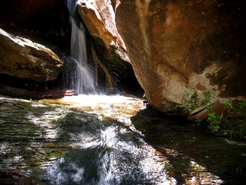 A serene waterfall cascades between large rocks, creating ripples in a tranquil pool below. Sunlight glimmers on the water.