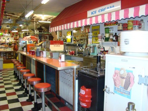 A retro ice cream parlor with a checkered floor, colorful decor, and a long counter with red stools.