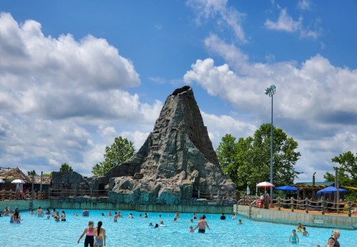 A large water park pool with a rocky mountain feature, filled with people enjoying the sunny day under a blue sky.