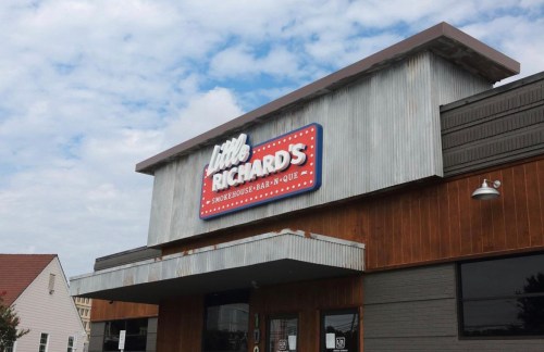 Sign for "Little Richard's Smokehouse Bar-B-Que" on a building with a rustic exterior and cloudy sky above.