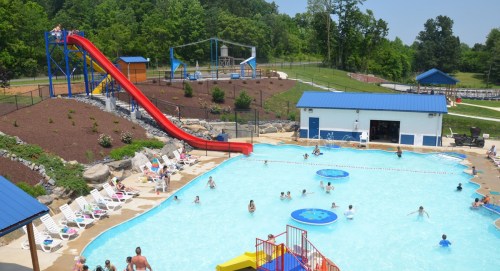 A vibrant pool area with a water slide, sun loungers, and people swimming, surrounded by greenery and recreational facilities.