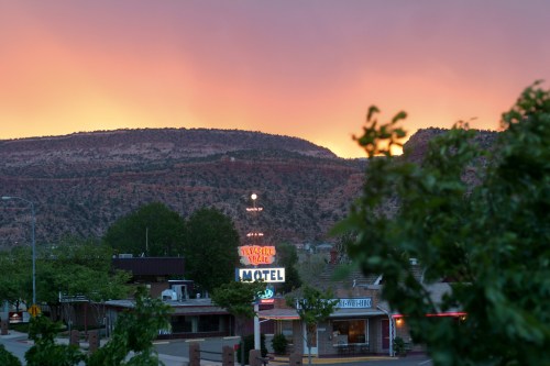 A sunset casts a warm glow over a motel and surrounding trees, with mountains in the background.