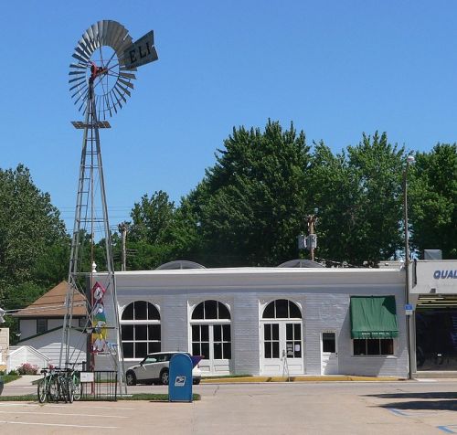 A windmill stands beside a white building with green awnings, surrounded by trees and a blue mailbox in the foreground.