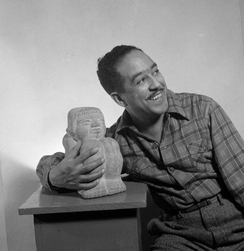 A smiling man in a plaid shirt holds a stone sculpture while sitting beside a small table.
