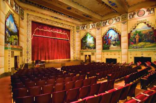 Interior of a vintage theater featuring a red curtain, ornate murals, and rows of empty seats.