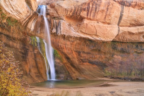 A serene waterfall cascades down a rocky cliff into a calm pool, surrounded by autumn foliage and rugged terrain.