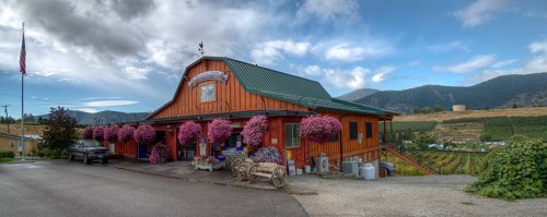 A rustic wooden building with hanging flower baskets, surrounded by vineyards and mountains under a cloudy sky.