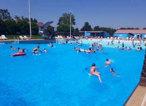 A busy outdoor pool filled with people swimming and relaxing on floats under a clear blue sky.