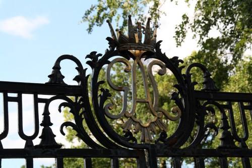Ornate wrought iron gate featuring a decorative crown and intricate design elements against a backdrop of greenery.