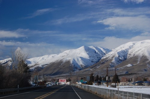 Snow-capped mountains rise against a blue sky, with a road and small buildings in the foreground.