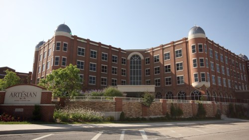 A large brick building with domed roofs, surrounded by greenery and a sign that reads "Artesian."