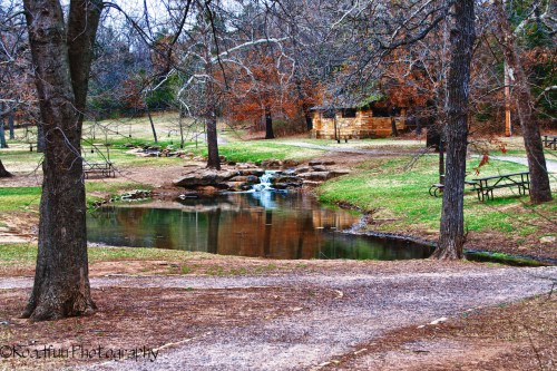 A serene park scene featuring a small stream, trees, and a picnic area with a rustic building in the background.