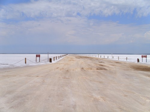A wide, sandy path leads to a vast, white salt flat under a cloudy sky, with signs on either side.