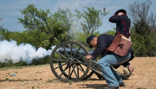 Two soldiers operate a cannon, with smoke billowing from the barrel, set against a grassy landscape.
