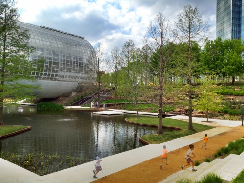 Children play near a serene pond surrounded by greenery and a modern glass building in a park setting.