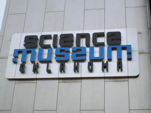 Sign for the Science Museum Oklahoma, featuring bold, modern lettering in blue and black on a light background.