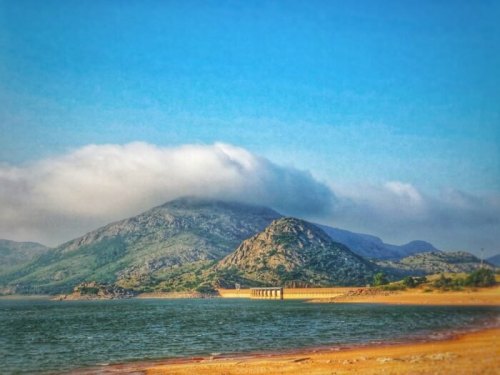A serene landscape featuring mountains, a calm lake, and a cloudy sky, with sandy shores in the foreground.