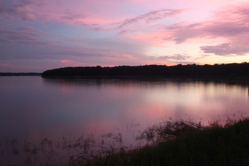 A serene lake at sunset, reflecting pink and purple hues in the water, surrounded by trees and gentle clouds.