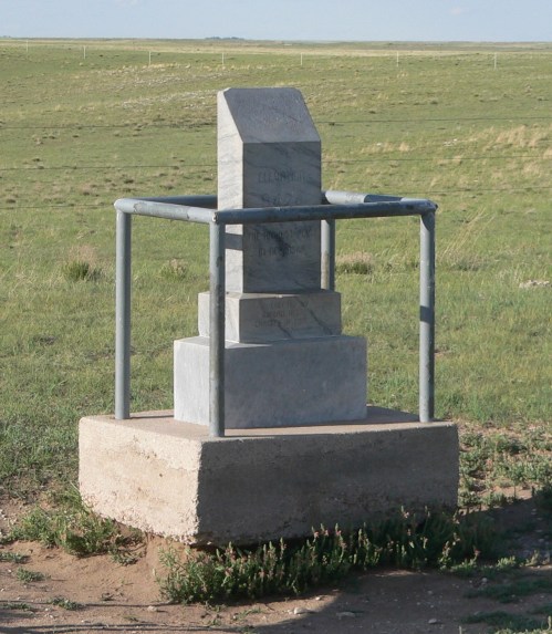 A stone monument surrounded by a metal frame, set on a concrete base in a grassy, open landscape.