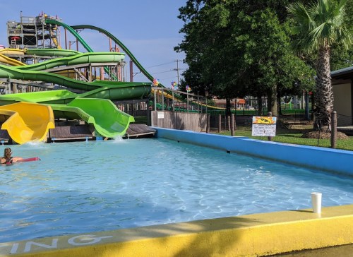 A water park scene featuring colorful slides, a pool, and a person swimming. Palm trees are visible in the background.