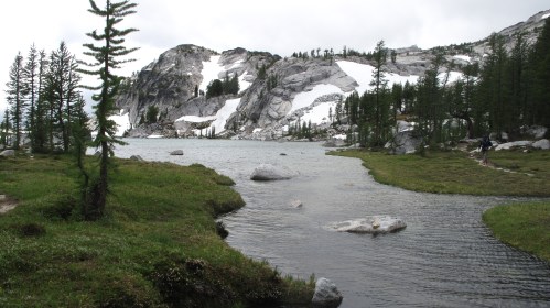 A serene mountain landscape featuring a lake, rocky cliffs, and patches of snow, surrounded by lush greenery and trees.