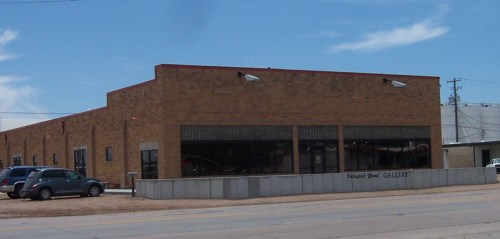 A brick building with large windows, labeled "Gallery," located on a street with parked cars nearby.