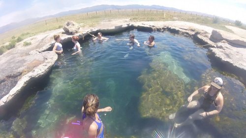 A group of people enjoying a natural hot spring surrounded by rocks and greenery under a clear blue sky.
