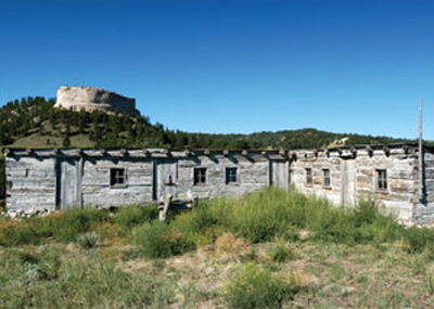 A weathered wooden building surrounded by grass, with a rocky hill in the background under a clear blue sky.