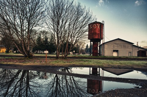 A red water tower and a building reflected in a puddle, surrounded by bare trees and a cloudy sky.