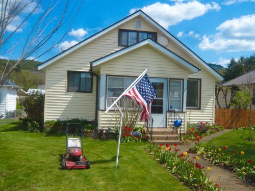 A yellow house with a front lawn, American flag, and a lawn mower beside colorful flower beds under a blue sky.