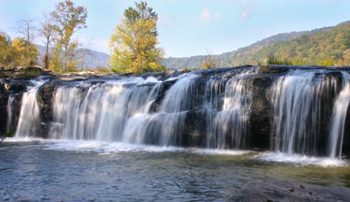 A serene waterfall cascades over rocks, surrounded by trees and mountains under a clear blue sky.