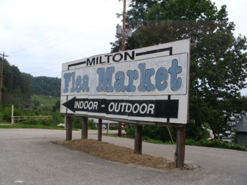 Sign for Milton Flea Market, indicating indoor and outdoor shopping options, surrounded by greenery.