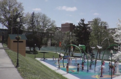 Children playing in a splash pad at a park, with trees and buildings in the background under a clear blue sky.