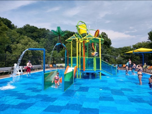 A colorful water play area with slides and fountains in a pool, surrounded by people enjoying the water on a sunny day.