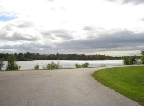 A serene lakeside view with a winding path, lush greenery, and cloudy skies in the background.