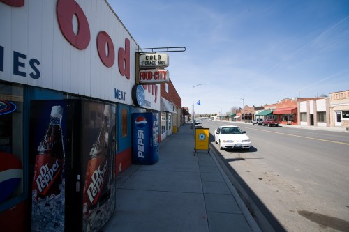 A street view of a small town featuring a grocery store, parked car, and storefronts under a clear blue sky.
