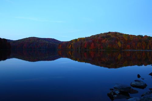 A serene lake reflecting colorful autumn trees and hills under a clear blue sky at dusk.