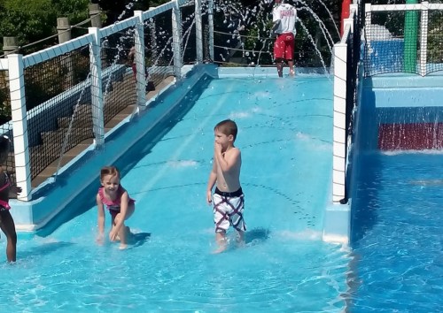 Two children play in a water slide area, splashing in the pool while water sprays around them.
