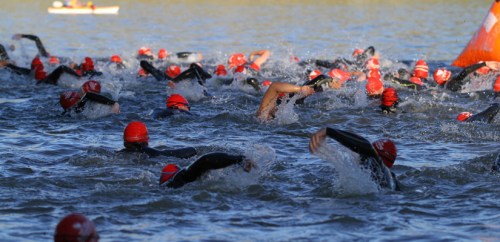A large group of swimmers in red caps competing in open water, creating splashes as they swim.