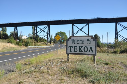 A welcome sign for Tekoa beside a winding road, with a railway trestle bridge in the background under a clear blue sky.