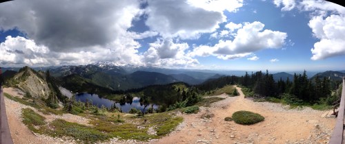 A panoramic view of mountains, a lake, and a blue sky with clouds, surrounded by lush greenery and rocky terrain.