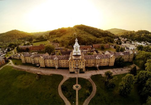 Aerial view of a historic building surrounded by greenery and hills, with a sunset in the background.