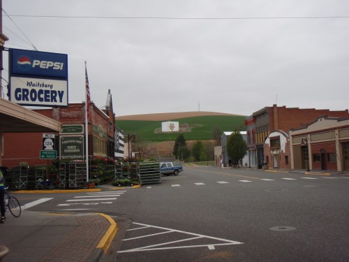 A small town street with grocery stores, a Pepsi sign, and green hills in the background under a cloudy sky.