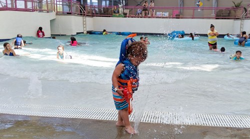 A child in a life jacket plays with water at a pool, surrounded by other kids enjoying the water.