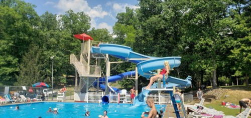 A busy pool area with a large water slide, people swimming, and sunbathing under a clear blue sky.