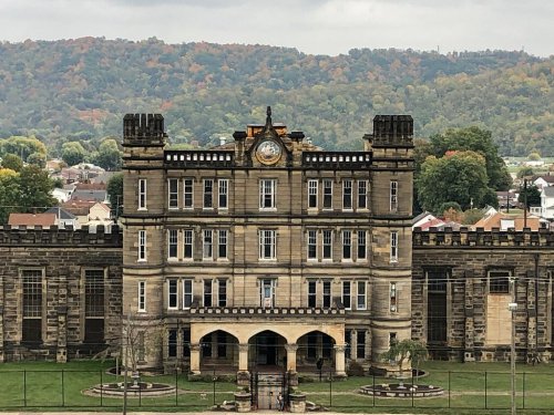 Historic stone building with towers and a clock, set against a backdrop of colorful autumn trees and hills.